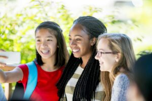 A multi-ethnic group of teenage girls are smiling and taking a selfie together on their cell phones.