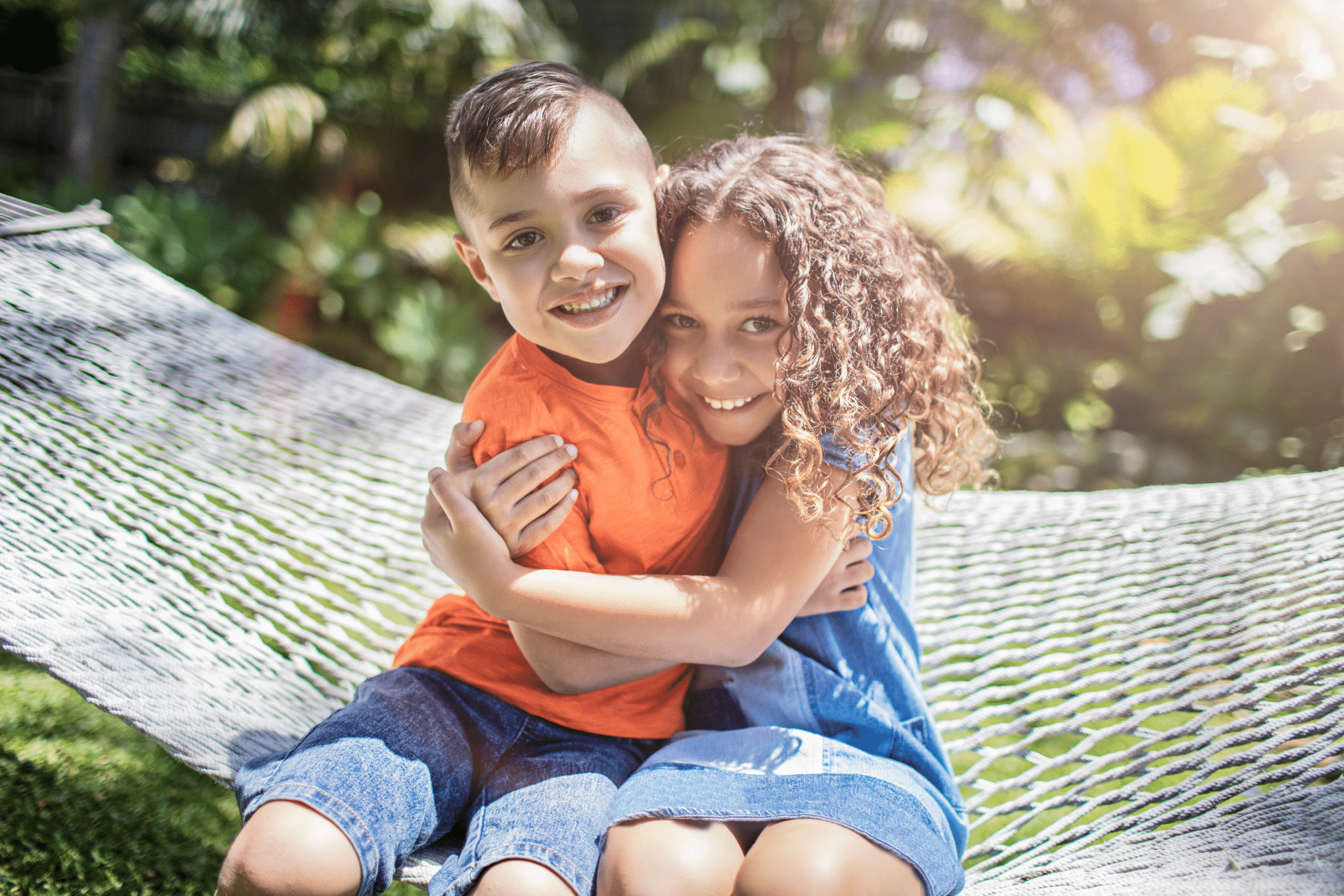 Two smiling children hugging while sitting together on a hammock outdoors